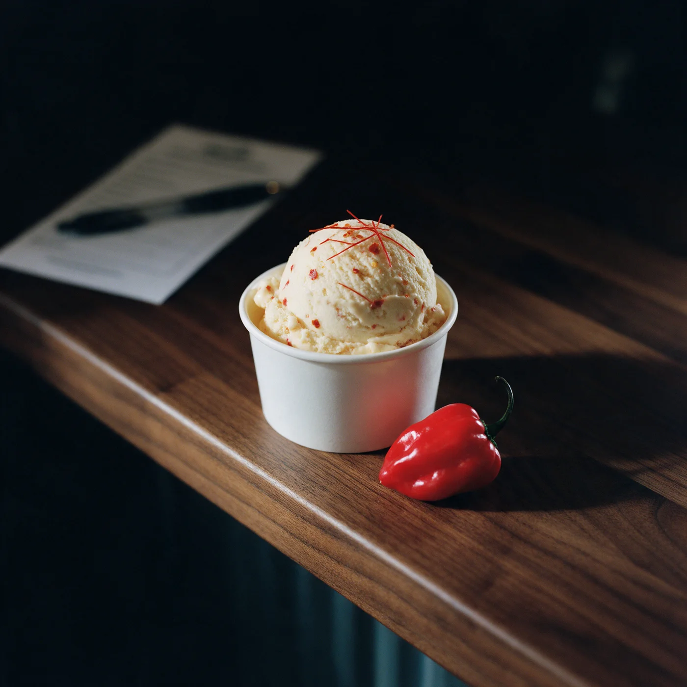 Styled concept photograph: a scoop of pale ice cream flecked with red chili in a white cup, a habanero beside it, a waiver form and pen blurred behind