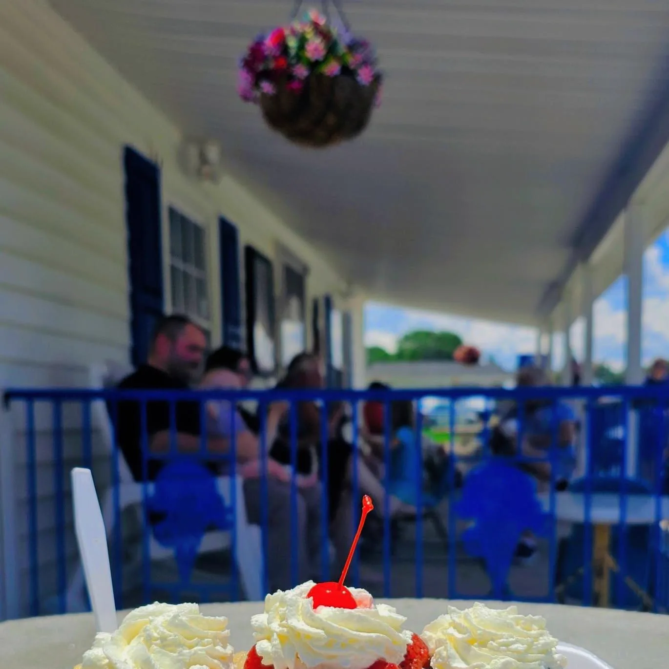 A strawberry shortcake sundae with whipped cream and a cherry, with the porch crowd out of focus behind it